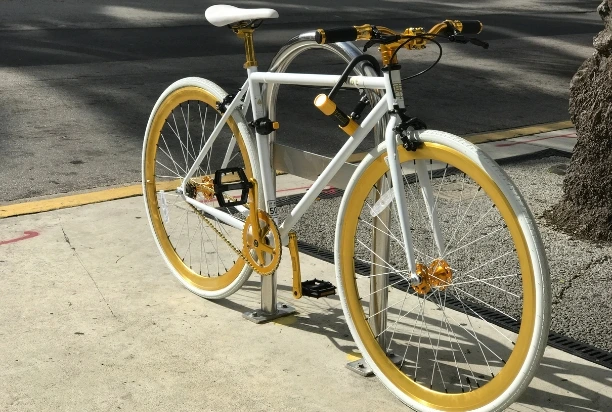 Ein weißes Fahrrad mit goldenen und gelben Farbakzenten, das in einer Straße an einem Fahrradständer angeschlossen ist.